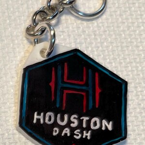 May include: A keychain with a black and white hexagonal pendant. The pendant has the text "HOUSTON DASH" in white, a red and blue design with the letter "H", and a silver chain and ring. The keychain is a sports accessory.
