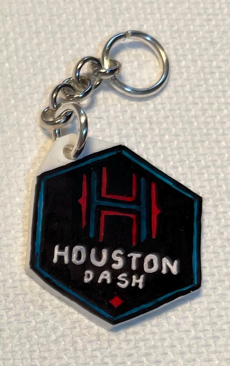 May include: A keychain with a black and white hexagonal pendant. The pendant has the text "HOUSTON DASH" in white, a red and blue design with the letter "H", and a silver chain and ring. The keychain is a sports accessory.