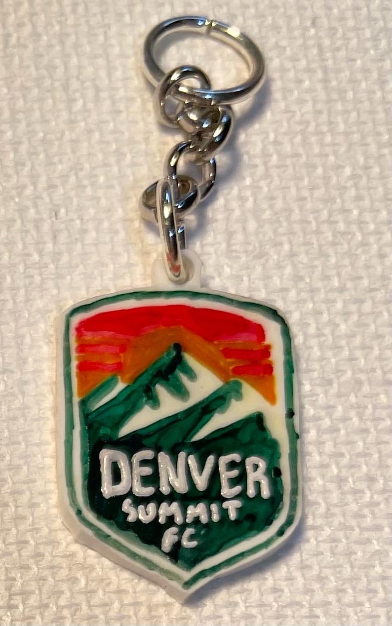 May include: A keychain with a rectangular pendant depicting a mountain scene in green, orange, and red. The text "DENVER SUMMIT FC" is in white. The keychain has a silver chain and ring.