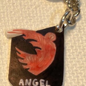 May include: A keychain with a black shield design, featuring a red and orange bird emblem and the text "ANGEL CITY FC" in white. It has a silver-coloured ring and chain.