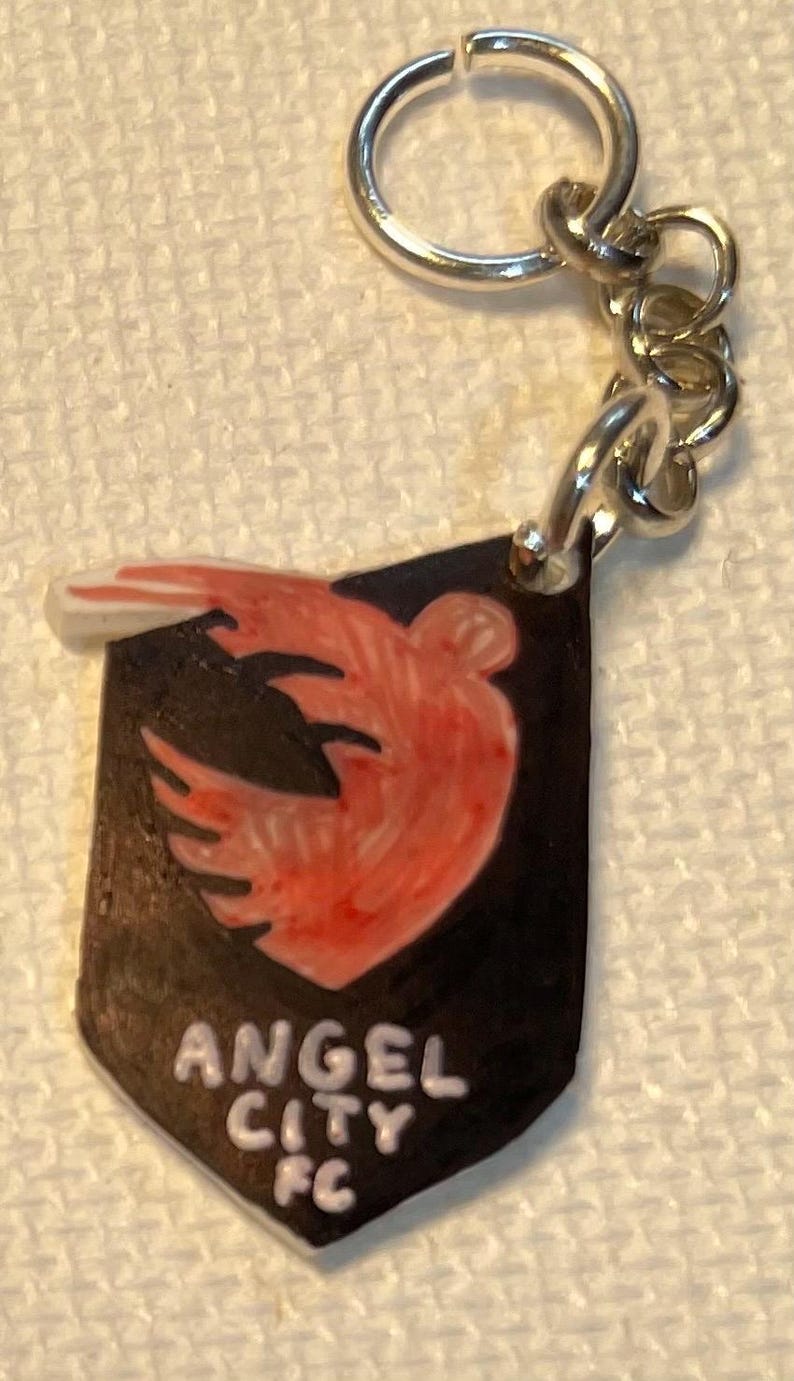 May include: A keychain with a black shield design, featuring a red and orange bird emblem and the text "ANGEL CITY FC" in white. It has a silver-coloured ring and chain.