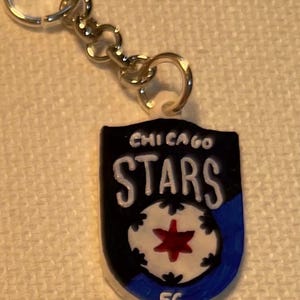 May include: A keychain with a shield-shaped emblem. The emblem displays the text "CHICAGO STARS" in white on a black background. It features a white soccer ball design with a red star and a blue lower edge.