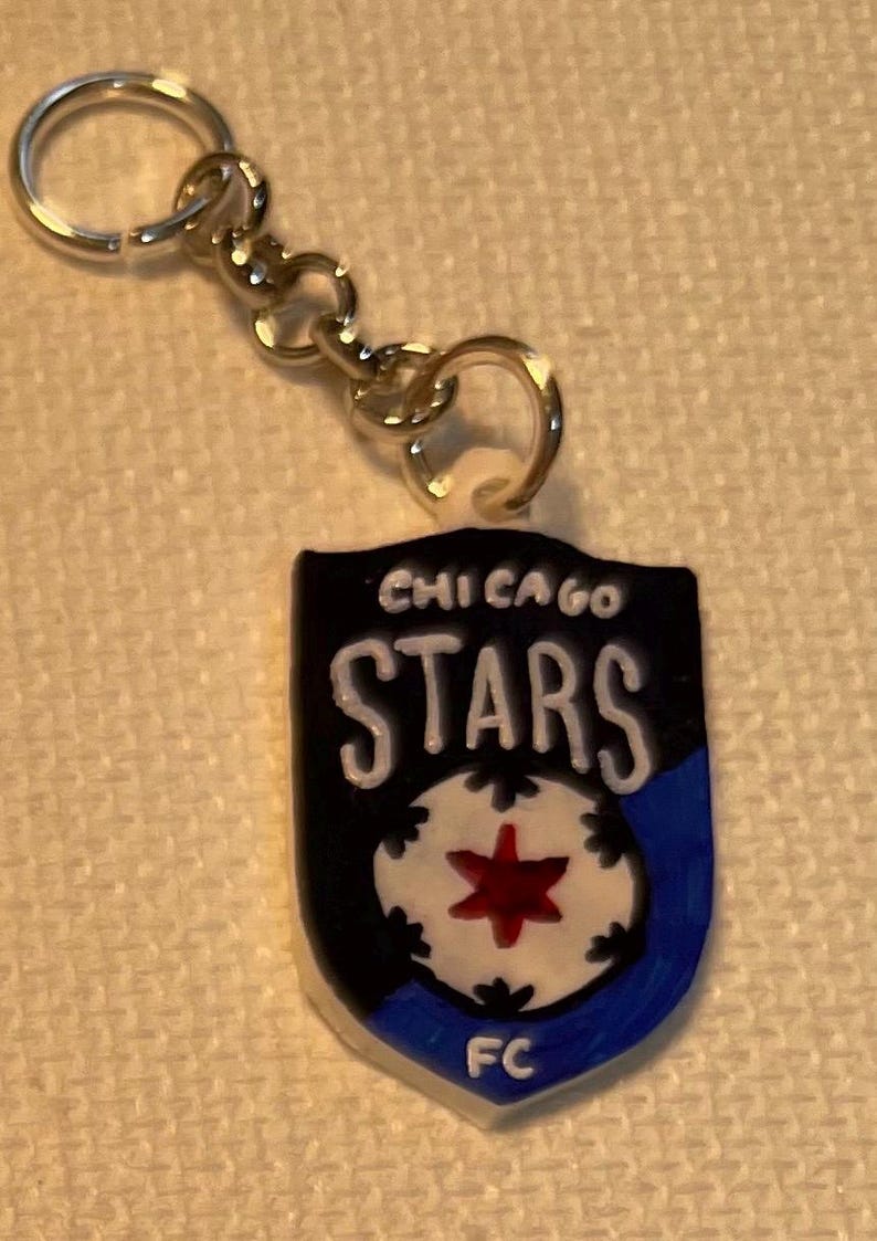 May include: A keychain with a shield-shaped emblem. The emblem displays the text "CHICAGO STARS" in white on a black background. It features a white soccer ball design with a red star and a blue lower edge.