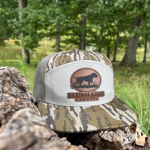 Puede incluir: Gorra de camionero con estampado de camuflaje y panel frontal beige. El panel frontal presenta un parche de cuero marrón con la silueta de un perro y el texto "FLUSH LINE APPAREL". La gorra tiene una parte trasera de malla y una visera curva.