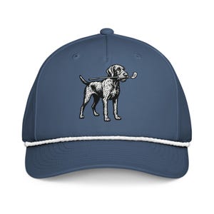 以下が含まれることがあります： 白ロープのディテールと、ゴルフのクラブをくわえた犬の白黒イラストが特徴の、ネイビーブルーの野球帽。帽子はカーブしたつばと上部にボタンが付いています。