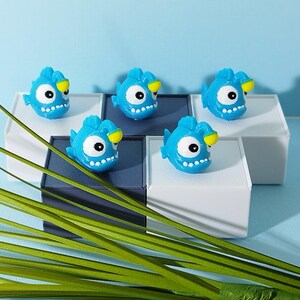 Puede incluir: Cinco juguetes azules con forma de pez, con ojos blancos y aletas amarillas, están dispuestos sobre bloques blancos y azul oscuro. Los juguetes tienen un diseño de dibujos animados con pequeños dientes blancos. Hojas verdes en primer plano sobre fondo azul claro.