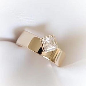 May include: A gold ring with a square-cut diamond. The ring has a thick band and a bezel setting that holds the diamond securely. The diamond is clear and reflects light, creating a sparkling effect. The ring is a piece of jewelry.