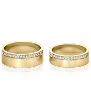 Puede incluir: Dos anillos de boda dorados con una fila de pequeñas piedras preciosas transparentes incrustadas en el borde superior. Los anillos se muestran sobre un fondo blanco.