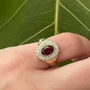 Könnte beinhalten: Ein Goldring mit einem oval geschliffenen, tiefroten Edelstein, der von einem Kranz aus kleinen weißen Steinen umgeben ist. An den Fingern befinden sich weitere Goldringe, darunter einer mit einer Reihe kleiner grüner Steine und ein strukturierter Ring.
