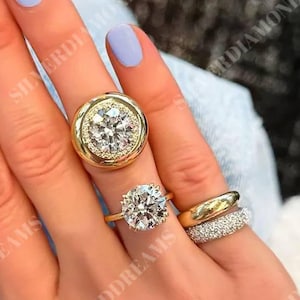 Op de afbeelding: Een verzameling van drie ringen gedragen aan de vingers. Eén ring heeft een grote ronde briljant geslepen middensteen omringd door een halo van kleinere stenen in een gouden zetting. Een andere ring heeft een gezette ronde steen en de derde is een gouden band.