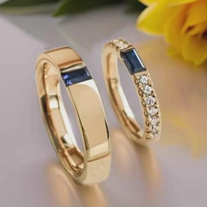 Può includere: Due anelli d'oro. Uno presenta una gemma blu rettangolare, l'altro una gemma blu simile e piccole gemme rotonde e trasparenti. Gli anelli sono ambientati su uno sfondo morbido e neutro con un fiore giallo.