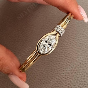 Pode incluir: Uma pulseira de ouro com um diamante central de corte marquise engastado numa moldura de ouro lisa. O design inclui uma pequena peça circular decorativa incrustada com pequenos diamantes pavé posicionada sobre a pedra principal.