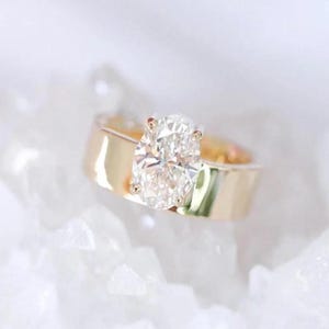 May include: A gold band ring with a large, oval-cut diamond. The diamond is set with small prongs, and the band has a wide, flat design. The ring is displayed on a white, textured surface.