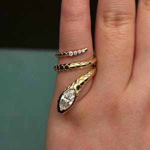 Puede incluir: Anillo de serpiente dorado con una gema en forma de diamante en la cabeza y pequeños diamantes en la cola. El anillo se enrolla alrededor del dedo en un diseño en espiral, creando una joya única y llamativa.