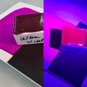 May include: A side-by-side comparison of a watercolor paint pan under normal light and under a black light. The pan is labeled "Ultra Violet" and the paint is a deep purple color under normal light and a bright pink color under black light.