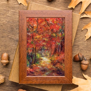 Peut inclure: Une peinture encadrée d'un chemin à travers une forêt avec des feuilles rouges, orange et jaunes. Le cadre est en bois brun et la peinture est sur une surface en bois avec des glands et des feuilles.