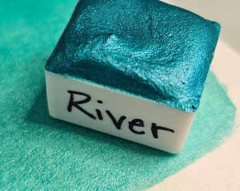 May include: A teal metallic watercolor paint pan with the word "River" written on the label.