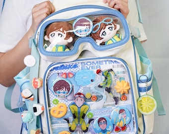 Puede incluir: Mochila blanca y azul con un bolsillo frontal transparente lleno de figuras y accesorios de dibujos animados. La mochila tiene ribetes y correas azules e incluye varios amuletos decorativos.
