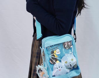 May include: Light blue crossbody bag with a clear front pocket displaying various charms and plush toys. The bag has a matching light blue strap and a cat-shaped keychain. The bag is worn over the shoulder.