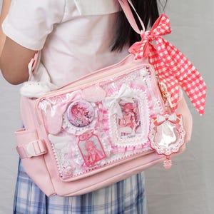 May include: A pink shoulder bag with a clear front pocket, decorated with badges, lace, and a red and white checkered ribbon. The bag has a plush bunny charm and a decorative keychain. The bag is designed with a kawaii aesthetic.