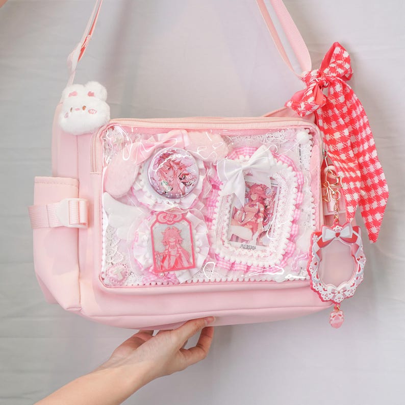 May include: A light pink shoulder bag with a clear front pocket filled with pink and white decorative items. The bag has a white plushie, a red and white checkered ribbon, and a keychain with a bow.