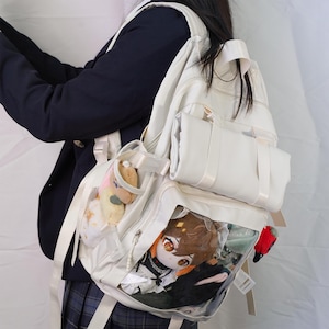 May include: Cream-coloured backpack with multiple compartments and clear pockets displaying plush toys. The backpack has white straps and a small handle. A person is wearing a navy blue blazer and a plaid skirt.