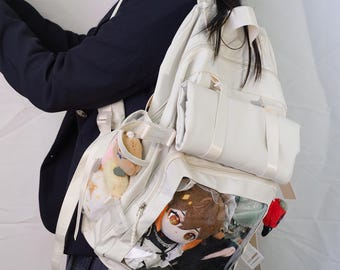 May include: Cream-coloured backpack with multiple compartments and clear pockets displaying plush toys. The backpack has white straps and a small handle. A person is wearing a navy blue blazer and a plaid skirt.