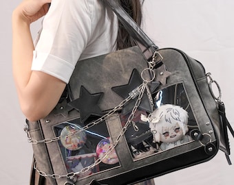 Könnte beinhalten: Graue Umhängetasche mit schwarzen Sternakzenten und Kettenanhänger. Die Tasche hat transparente Fensterfächer mit bunten Anstecknadeln und Plüschfiguren. Eine silberne Kette und ein Sternanhänger hängen an der Tasche.
