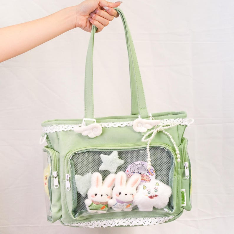 May include: A light green tote bag with a clear front pocket displaying plush toys, including two white bunny rabbits and a white cat. The bag has white lace trim, flower accents, and a pearl-like chain.