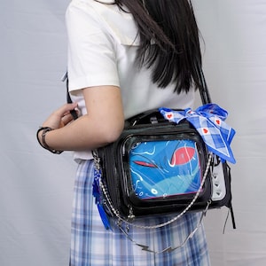 Könnte beinhalten: Schwarze Umhängetasche mit einem transparenten Frontpanel, das eine blaue Monstergrafik zeigt. Die Tasche hat einen silbernen Kettenriemen, einen blauen Federanhänger und einen blau-weiß karierten Schal. Die Tasche wird über einem weißen Hemd und einem blauen karierten Rock getragen.