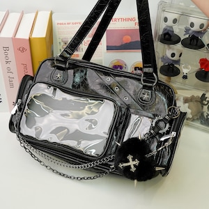 May include: Black faux leather handbag with a textured finish, featuring clear pockets and silver-tone hardware. Includes a chain strap, star accents, and a fluffy black charm with a cross. The bag has a top handle and a detachable coin purse.