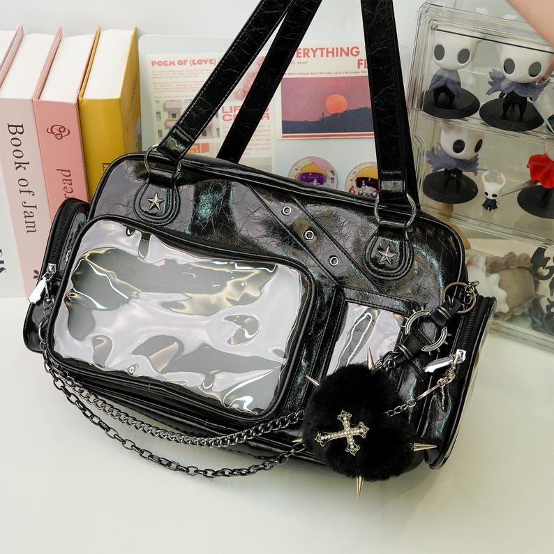 May include: Black faux leather handbag with a textured finish, featuring clear pockets and silver-tone hardware. Includes a chain strap, star accents, and a fluffy black charm with a cross. The bag has a top handle and a detachable coin purse.