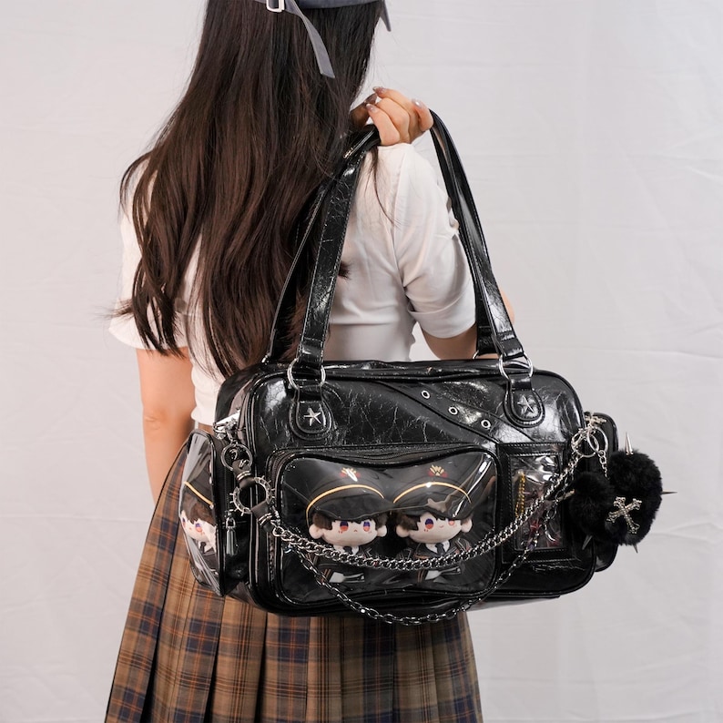 May include: Black faux leather handbag with multiple compartments, silver hardware, and a chain detail. The bag features clear pockets with plush dolls and a black pom-pom with a cross charm. The bag is carried by a person.