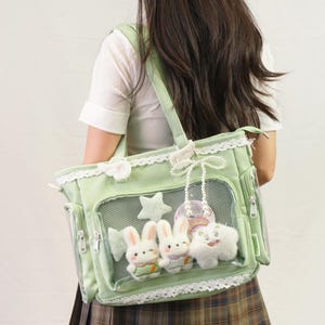 May include: A light green tote bag with a clear front panel, featuring plush bunny and star decorations. The bag has white lace trim and a matching shoulder strap. Additional small pockets and a zipper are visible.