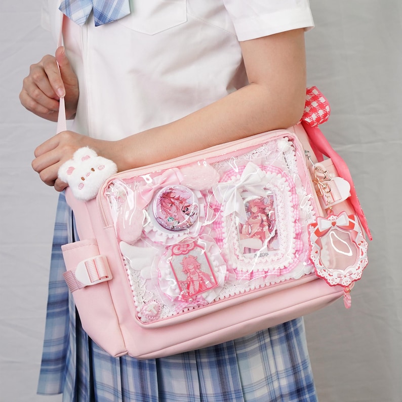 May include: A pink messenger bag with a clear front panel displaying decorative elements. The bag features a white plush bunny charm, a pink bow, and various pink and white embellishments. The bag has a shoulder strap and a buckle closure.