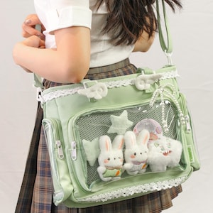 May include: A light green messenger bag with a clear front pocket displaying plush bunny and cat toys. The bag has white lace trim, a shoulder strap, and decorative charms.