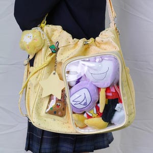 May include: A yellow shoulder bag with a star pattern and a clear pocket filled with plush toys. The bag has a drawstring closure and a long strap. Additional charms include a star, a cookie, and a fluffy yellow charm.