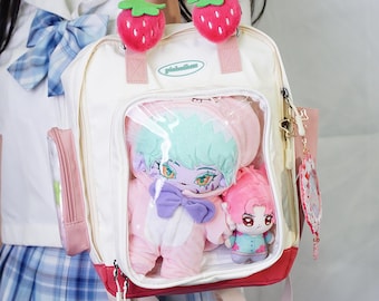 Könnte beinhalten: Ein weiß-rosa Rucksack mit transparentem Frontpanel, gefüllt mit Plüschpuppen. Der Rucksack hat Erdbeer-Anhänger an den Griffen. Eine kleine rosa Seitentasche und ein roter Boden vervollständigen das Design.