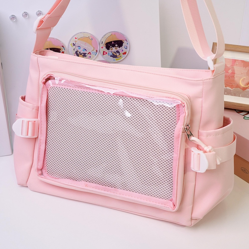 May include: A light pink messenger bag with a clear mesh pocket on the front. The bag has a shoulder strap, side pockets, and white plastic buckles. Three round pins with cartoon characters are visible.