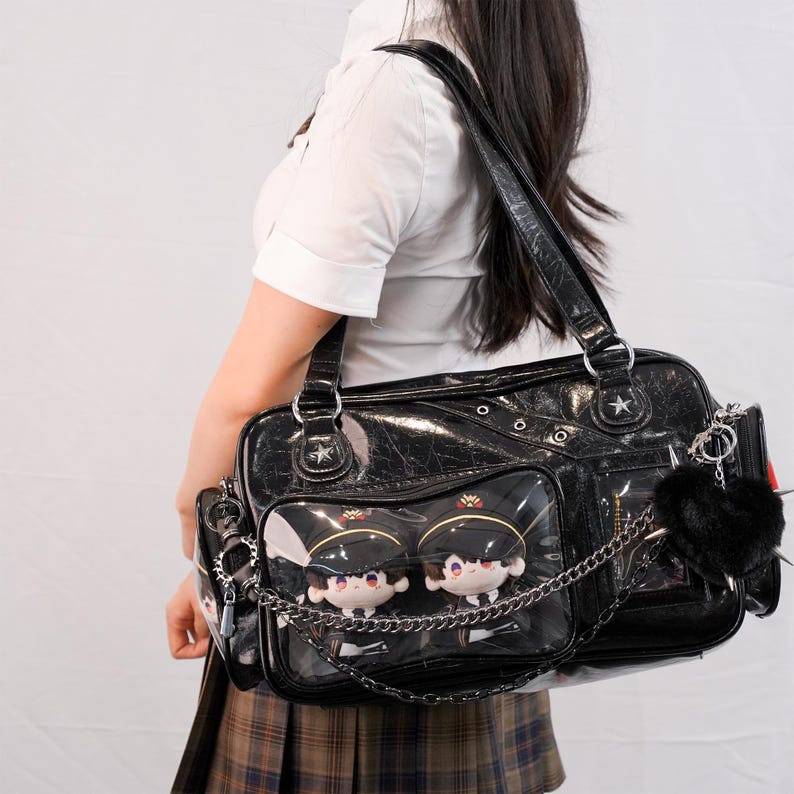 May include: Black patent leather shoulder bag with multiple compartments and silver-tone hardware. The bag features clear pockets displaying plush dolls and a chain detail. A black pom-pom and keychains hang from the side.