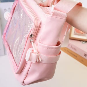 May include: A light pink crossbody bag with a clear front panel and a mesh pocket. The bag has a zipper closure, a small side pocket, and a white strap. The bag is made of a soft, durable material.