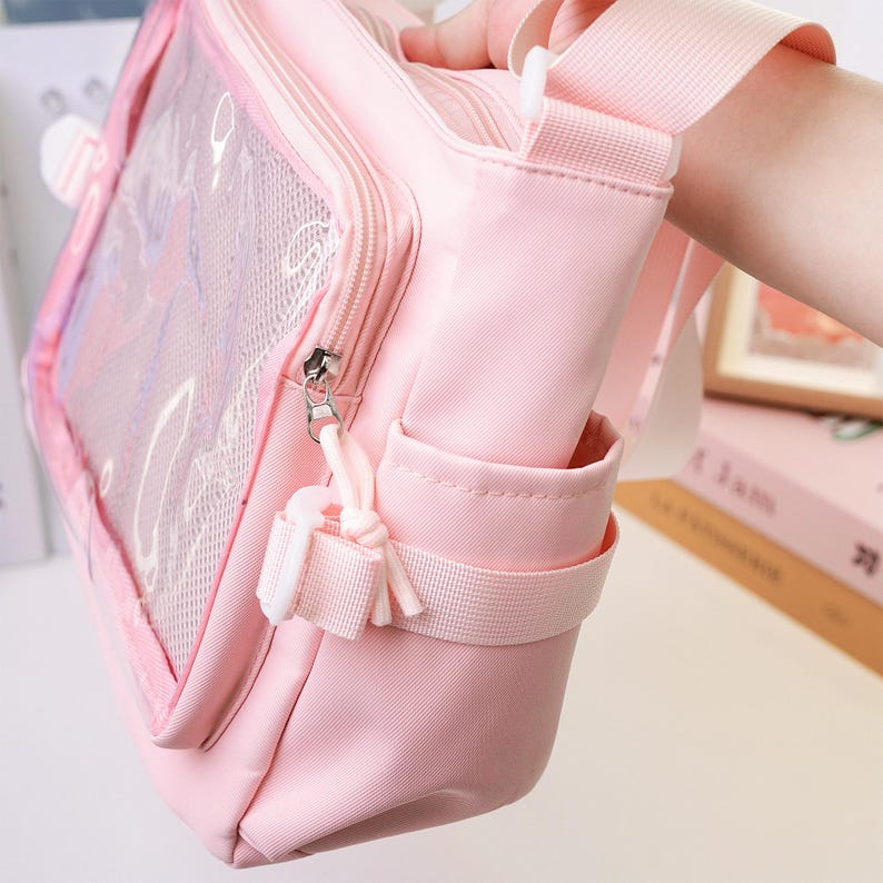 May include: A light pink crossbody bag with a clear front panel and a mesh pocket. The bag has a zipper closure, a small side pocket, and a white strap. The bag is made of a soft, durable material.