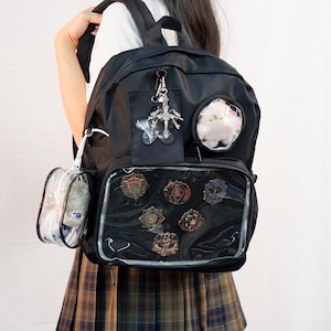 May include: Black backpack with multiple clear pockets displaying decorative pins and a plush toy. The backpack has a silver charm and a small clear pouch attached. The bag is worn by a person wearing a plaid skirt.