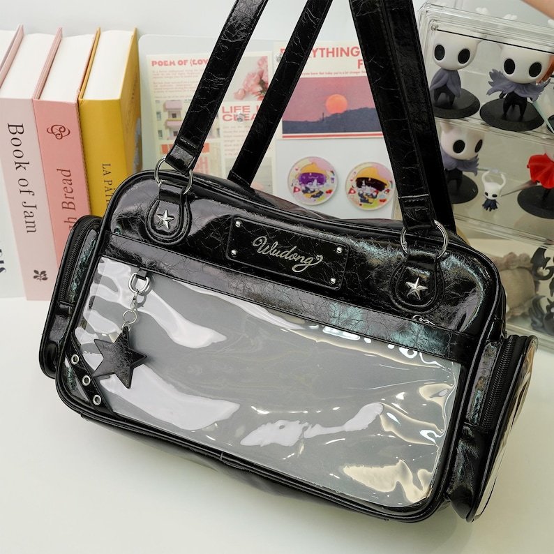 May include: Black patent leather handbag with a clear front panel. The bag features a star-shaped charm, silver hardware, and the word "Wudong" on the front. The bag has a top handle and a shoulder strap.