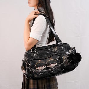 May include: Black patent leather shoulder bag with star accents and clear pockets displaying plush dolls. The bag features a chain and a black pom-pom charm. The bag has a top handle and a shoulder strap.