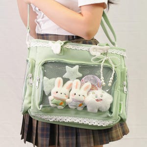 May include: A light green shoulder bag with a clear front pocket displaying plush bunny and star decorations. The bag has lace trim, multiple pockets, and a white key-shaped charm. The bag is carried by a light green strap.
