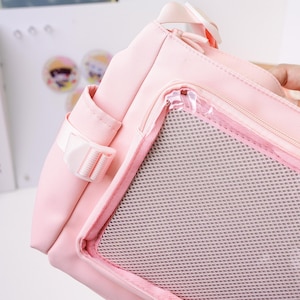 May include: A light pink tote bag with a mesh pocket and a white buckle. The bag has a zipper closure and a small side pocket. The bag is made of a durable material and has a handle.