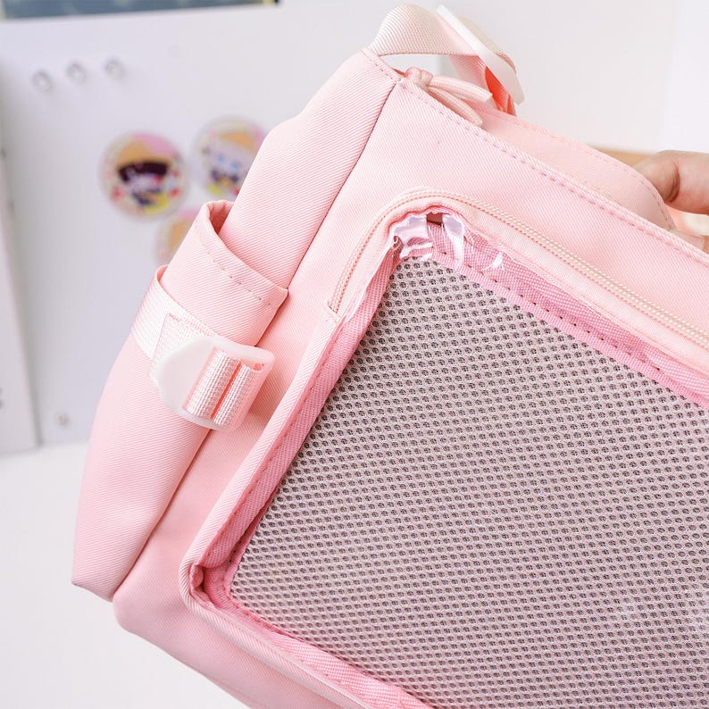 May include: A light pink tote bag with a mesh pocket and a white buckle. The bag has a zipper closure and a small side pocket. The bag is made of a durable material and has a handle.