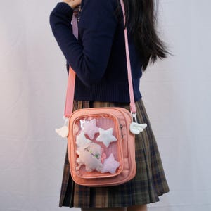 Puede incluir: Un bolso bandolera rosa con un bolsillo frontal transparente lleno de estrellas de peluche y otras formas. El bolso tiene una correa rosa y colgantes blancos con forma de ala. El bolso se lleva sobre un suéter azul oscuro y una falda a cuadros.