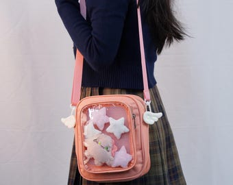 May include: A pink crossbody bag with a clear front pocket filled with plush stars and other shapes. The bag has a pink strap and white wing-shaped charms on the sides. The bag is worn over a dark blue sweater and plaid skirt.